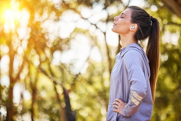 Woman in a lavender athletic top standing in a sunlit park with hands on hips, looking upward, wearing wireless earbuds.