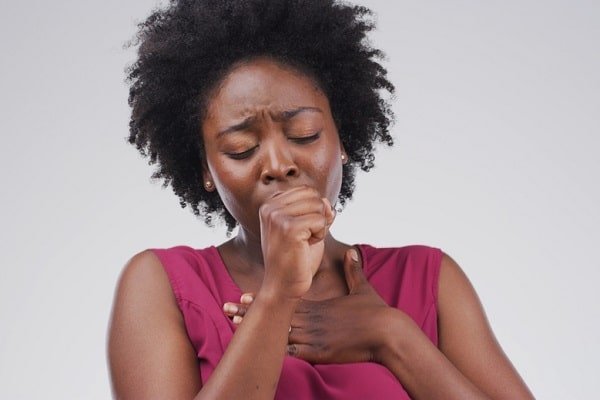 Black woman with natural hair coughing into her fist, wearing a pink sleeveless top.