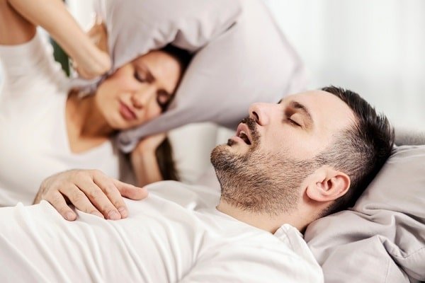 Man sleeping on a white bed while a woman behind him holds a pillow to her head, looking tired or overwhelmed.