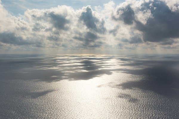 Sunlight glinting off a calm ocean under a cloudy sky, with a distant horizon.