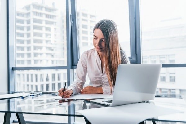 Woman using a laptop and writing on a notepad at a glass desk in a bright office with large windows behind her.