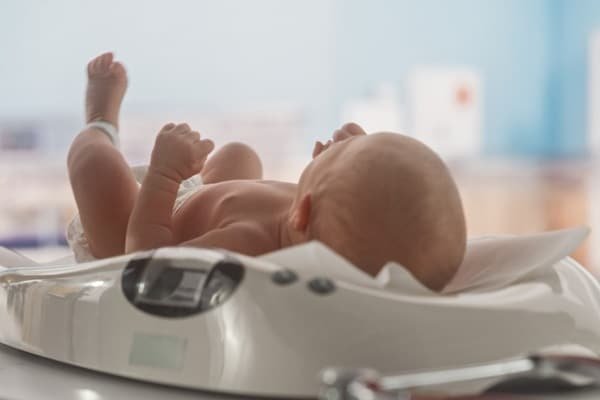 Newborn baby lying on a digital baby scale in a hospital setting, feet up for weighing.
