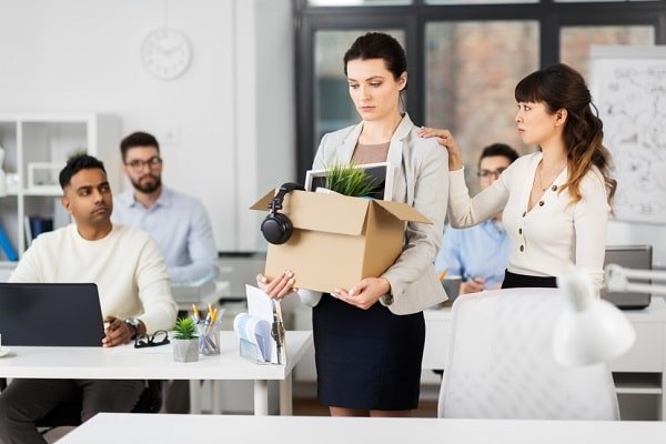 Businesswoman carries a box of personal belongings with a plant and headphones as coworkers look on in a bright open-plan office.