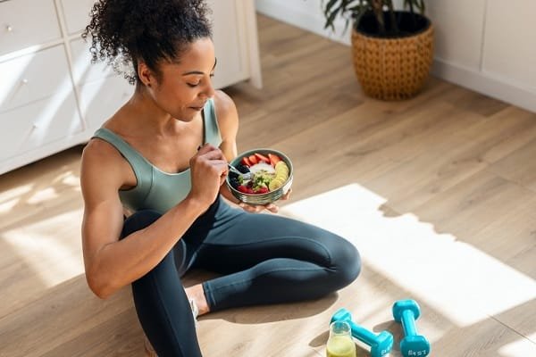 Woman in teal athletic wear sits on the floor and eats a colorful salad from a bowl after a home workout, with dumbbells nearby.