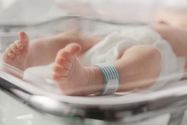 Newborn baby feet with a blue hospital identification band resting inside a clear neonatal bassinet/incubator.