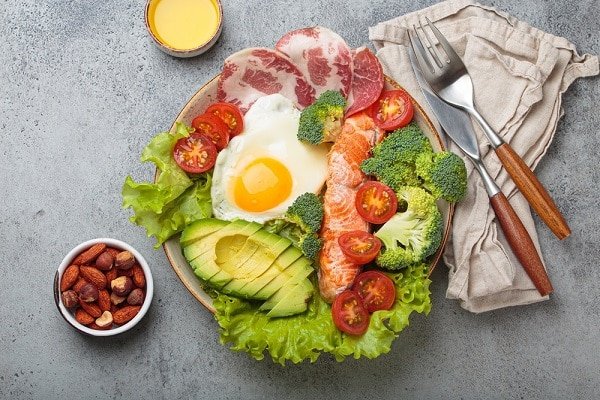 Breakfast plate with a sunny-side egg, avocado, broccoli, lettuce, cherry tomatoes and slices of bacon and smoked salmon.