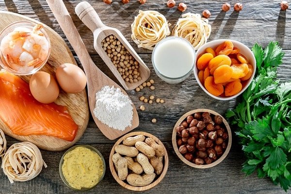 Top-down view of a pantry spread with salmon, eggs, pasta nests, flour, milk, chickpeas, dried apricots, nuts, and fresh parsley on a rustic wooden surface.