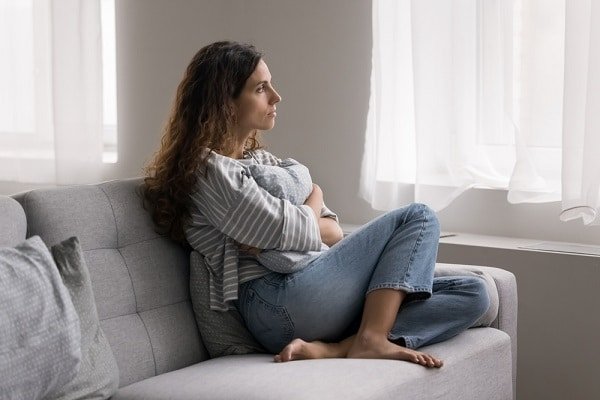 Woman seated on a gray sofa, hugging a pillow and looking out a bright window.