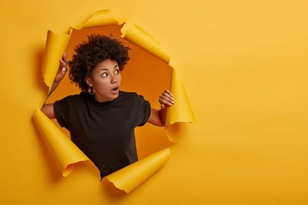 Woman peeking through a torn yellow paper backdrop with a surprised expression on her face.