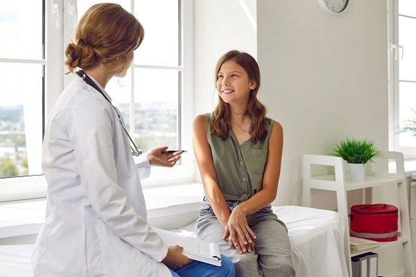 Doctor with stethoscope talking to a smiling patient seated on an exam bed in a bright clinic room by a window.