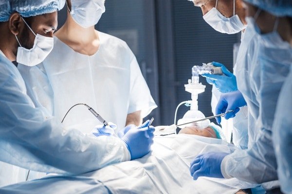 Surgeons in scrubs and masks perform surgery while an anesthesia provider manages the patient's airway with breathing equipment nearby.