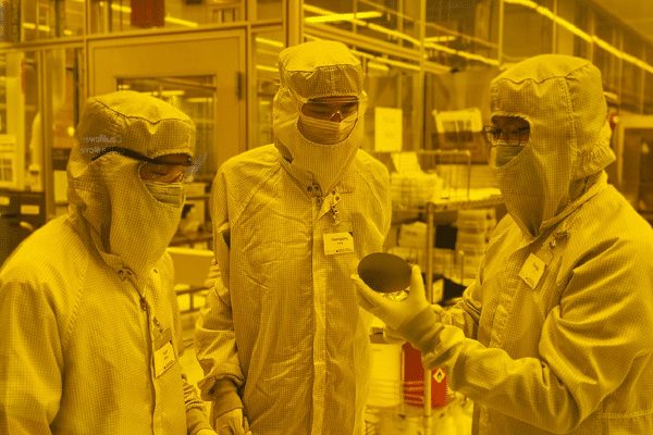Three workers in cleanroom suits and badges discuss in a high-tech manufacturing lab.