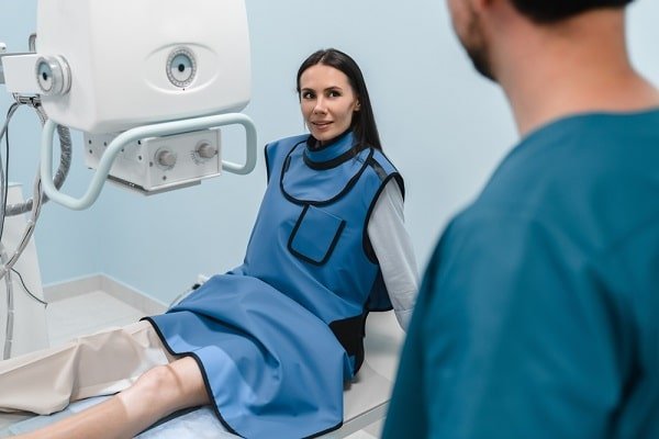 Patient sits on an examination table wearing a blue protective gown during an eye imaging procedure, with a large white imaging device adjacent and a clinician in teal scrubs standing nearby.