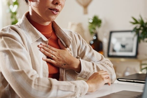 Person seated at a desk places a hand on their chest, wearing an orange turtleneck under a beige jacket, with a laptop and a framed photo in the background.