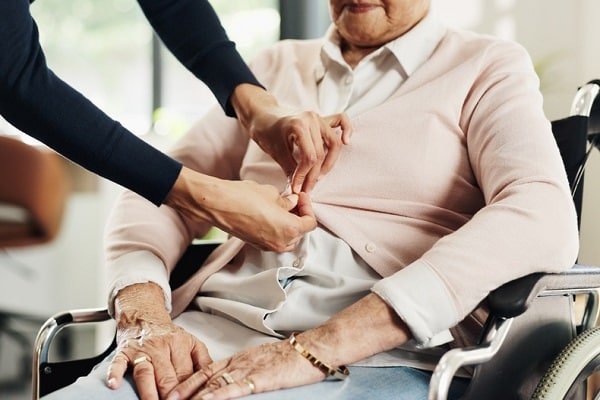 Caregiver helping an elderly woman in a wheelchair button her cardigan, providing gentle assistance.