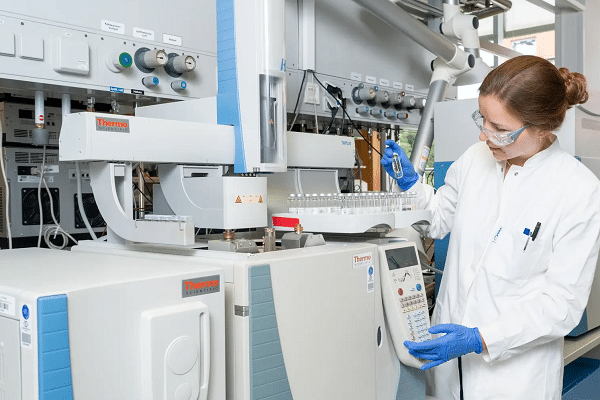 Female lab technician in safety goggles and blue gloves operates a Thermo Scientific chromatography/analytical instrument in a modern lab.