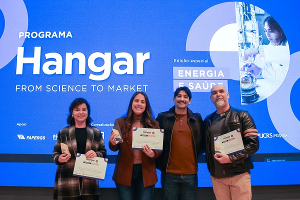 Four people stand in a row on a blue stage, each holding a certificate in front of a backdrop that says 'Hangar' and 'From Science to Market'.