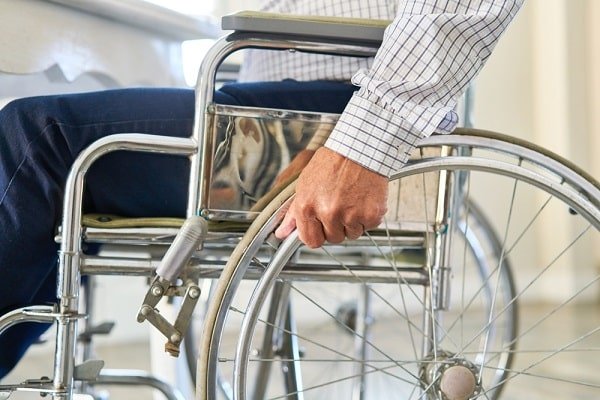 Person in a wheelchair gripping the wheel rim with a gloved-like hand area visible, illustrating manual wheelchair use in a clinical setting.
