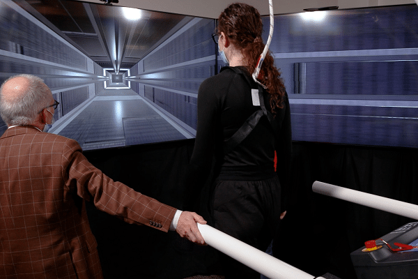 Older man in a brown jacket guides a person in black with a safety harness as they stand at a motion-simulation tunnel with a long corridor display on screens ahead.