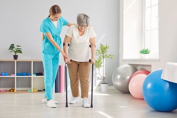 Caregiver helps an elderly woman walk with forearm crutches in a bright rehab room with exercise equipment nearby