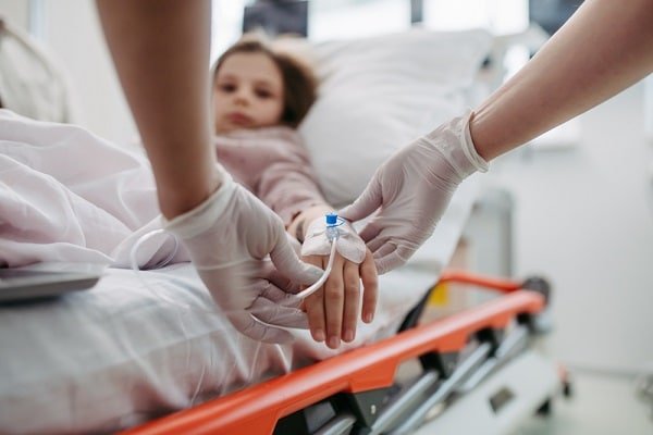 Child resting in a hospital bed while clinicians in gloves insert an IV line into the back of their hand.