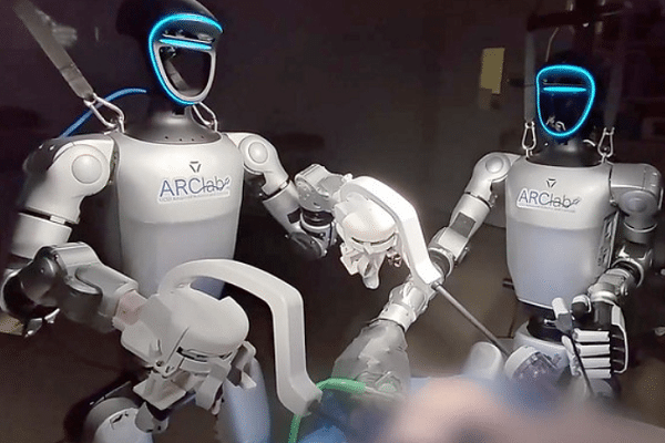 Two ARClab humanoid robots with blue illuminated faces assist a surgical robotic arm in a lab setting.