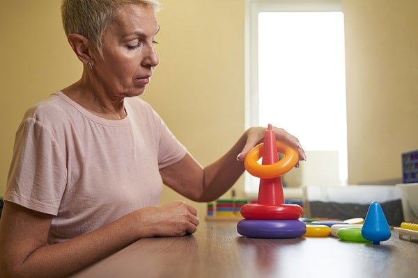 Older woman with short gray hair sits at a table, stacking colorful rings on a red cone toy, focused on playtime.