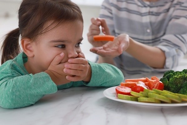 Child looks hesitant at mealtime while a parent offers carrot slices from a spoon, with a plate of vegetables nearby