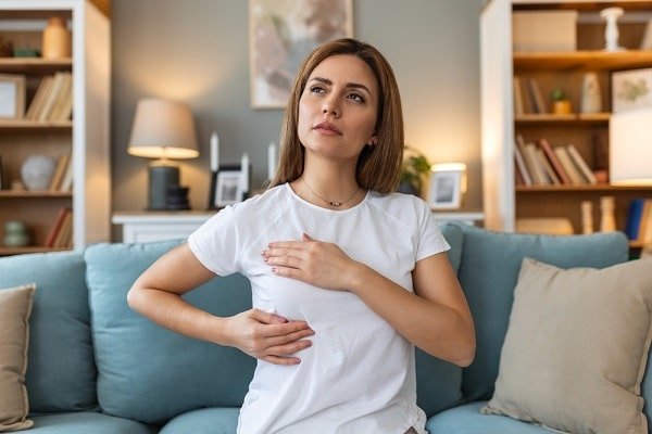Woman on a sofa clutches her chest and stomach, looking concerned, suggesting chest pain or abdominal discomfort in a living room setting.