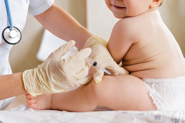 Healthcare professional in gloves giving a syringe injection to a baby on a changing table; stethoscope visible at left.