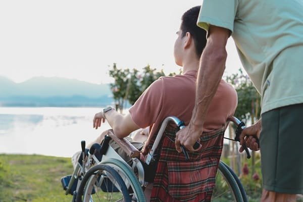 Person in a wheelchair being pushed along a grassy lakeside by another person outdoors.