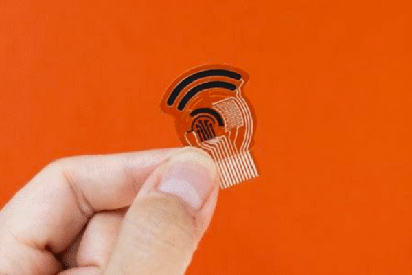 Close-up of a hand holding a small transparent NFC/RFID sticker with an antenna, about to be placed on a surface against an orange background.