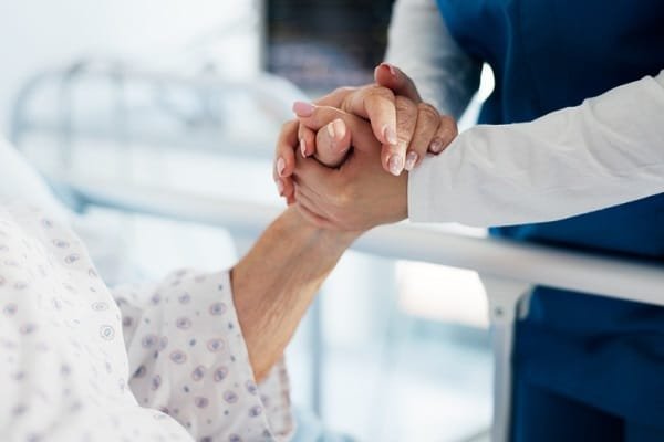 Nurse in blue scrubs holding hands with a patient in a hospital bed, offering comfort.