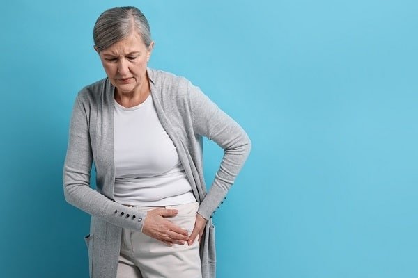 Older woman wearing a white shirt and gray cardigan clutches her abdomen with a pained expression against a blue background.