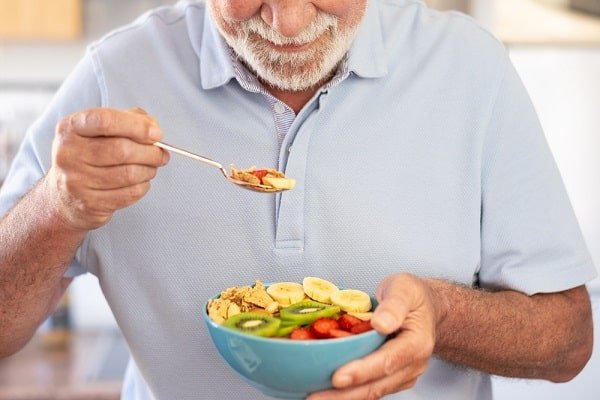 Senior man in a light blue polo eating a colorful fruit-and-vegetable salad from a blue bowl with a spoon nearby.