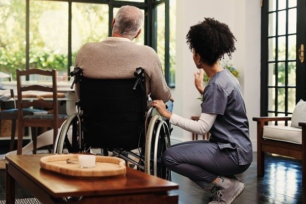 Caregiver kneels beside an elderly man in a wheelchair, in a bright living room, offering support and conversation.