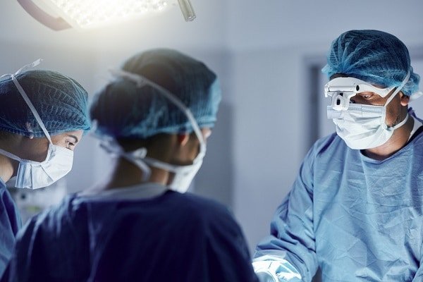 Surgeons in an operating room performing surgery under bright overhead lights and wearing masks, gowns, and caps.