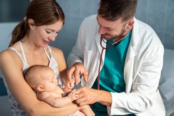 Mother cradling her baby while a doctor wearing scrubs and a white coat checks the infant's arm with a stethoscope nearby, in a clinic setting (pediatric checkup).