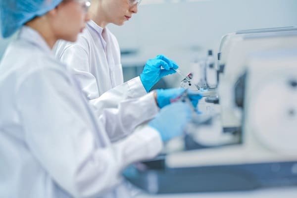 Two scientists in lab coats and blue gloves operate small vials at a benchtop instrument in a clean lab setting plus blurred equipment in the foreground of the photo.