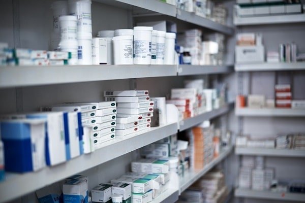 Pharmacy shelves stocked with bottles and boxes of medicines arranged in rows on gray shelves in a well-lit aisle.