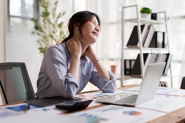 Woman at a messy desk in office, eyes closed, clutching her neck in fatigue or stress with a laptop and papers nearby.
