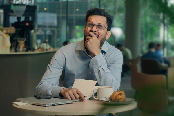 Man wearing glasses sits at a cafe table, resting his chin on his hand, with a laptop, notebook, coffee, and a pastry nearby.