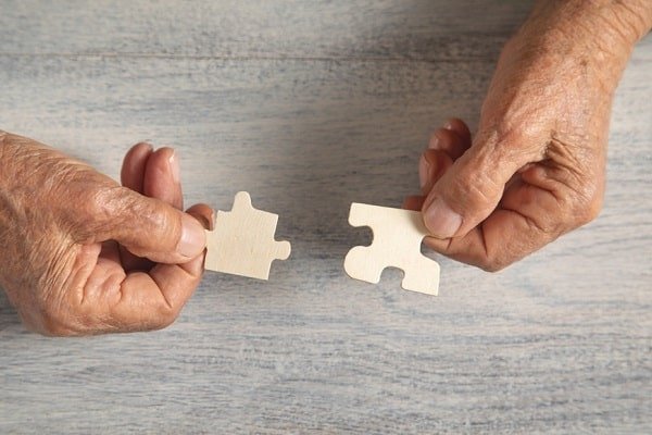 Two elderly hands hold puzzle pieces about to connect, symbolizing partnership and connection on a light wood surface and/or collaborative effort in aging.